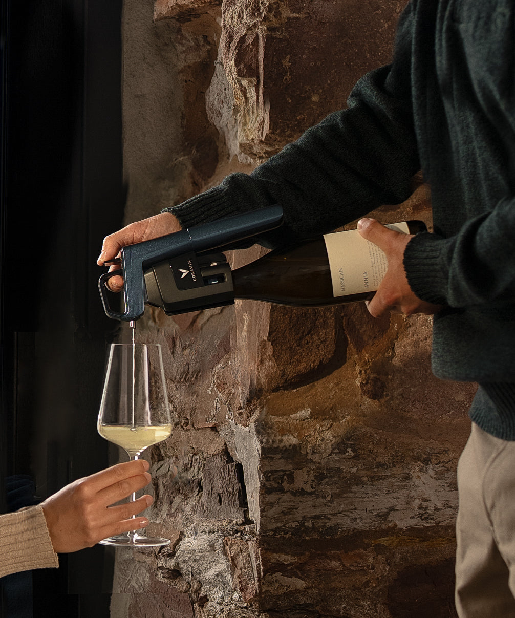 A person pours white wine from a bottle using Corvain Ocean Blue into a glass held by another person, set against a rustic stone wall background.