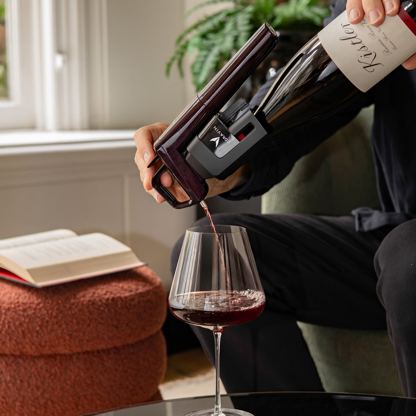 Person pouring red wine from a bottle into a glass, with a book and plant in the background.